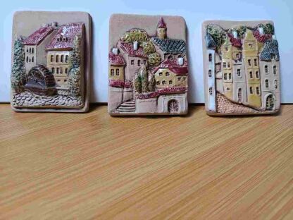 3 ceramiczne obrazki z zabudową miejską obok siebie w drobnych odstępach oparte o jasną ścianę 