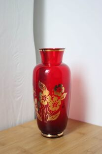 Wazon w kolorze czerwonego wina z roślinnym złotym wzorem i wąska szyjką na tle białej powierzchni
