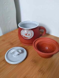 Ceramiczny kubek z leżącym obok ceglanym sitkiem i jasną pokrywką